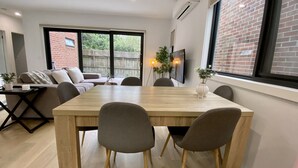 Een moderne eetruimte met een houten tafel en grijze stoelen, een bank en een aan de muur bevestigde televisie.