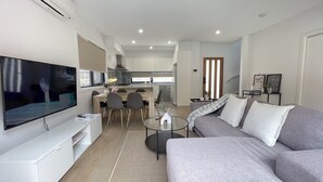 Een moderne woonkamer met een flatscreen televisie, een grijze bank en een eethoek met stoelen en een tafel.