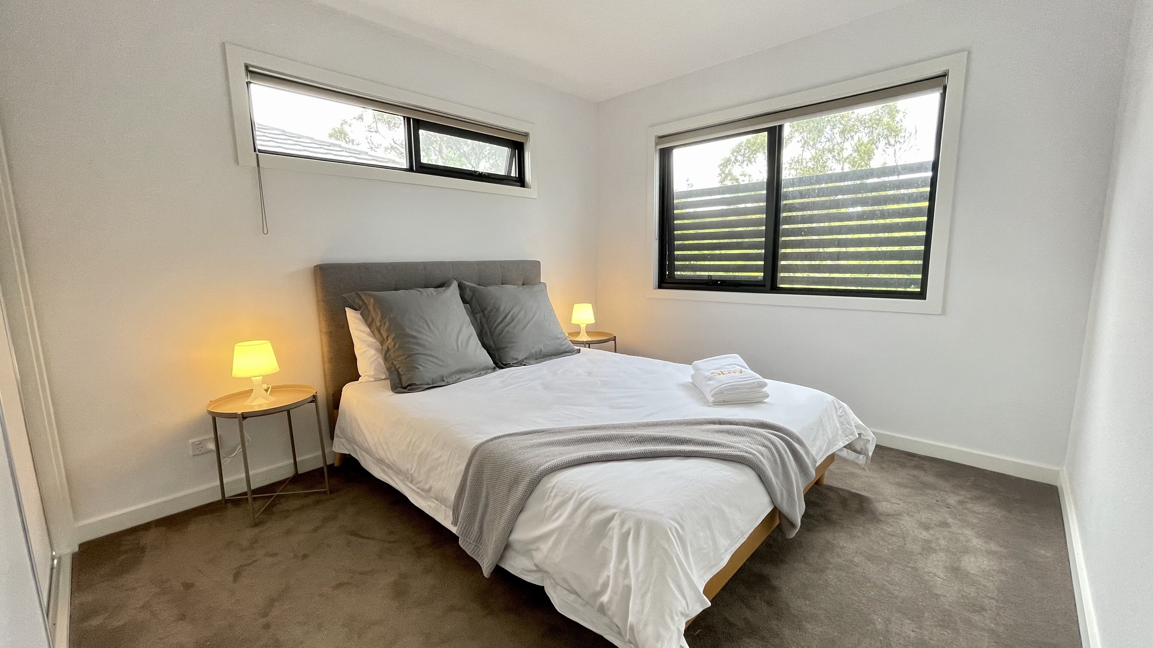 A bedroom with a bed, two bedside lamps, a window with blinds, and a small table with a lamp.