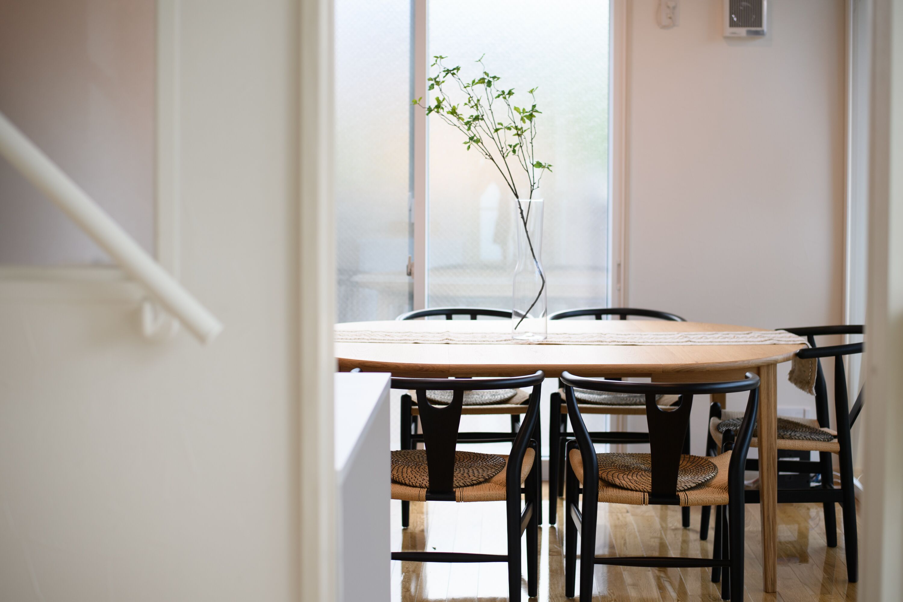 Uma área de refeições com uma mesa redonda, cadeiras pretas e um vaso com plantas verdes.