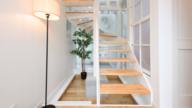 Un escalier moderne avec des marches en bois et une rampe en verre, une plante en pot et une lampe sur pied dans une pièce lumineuse dotée de grandes fenêtres.