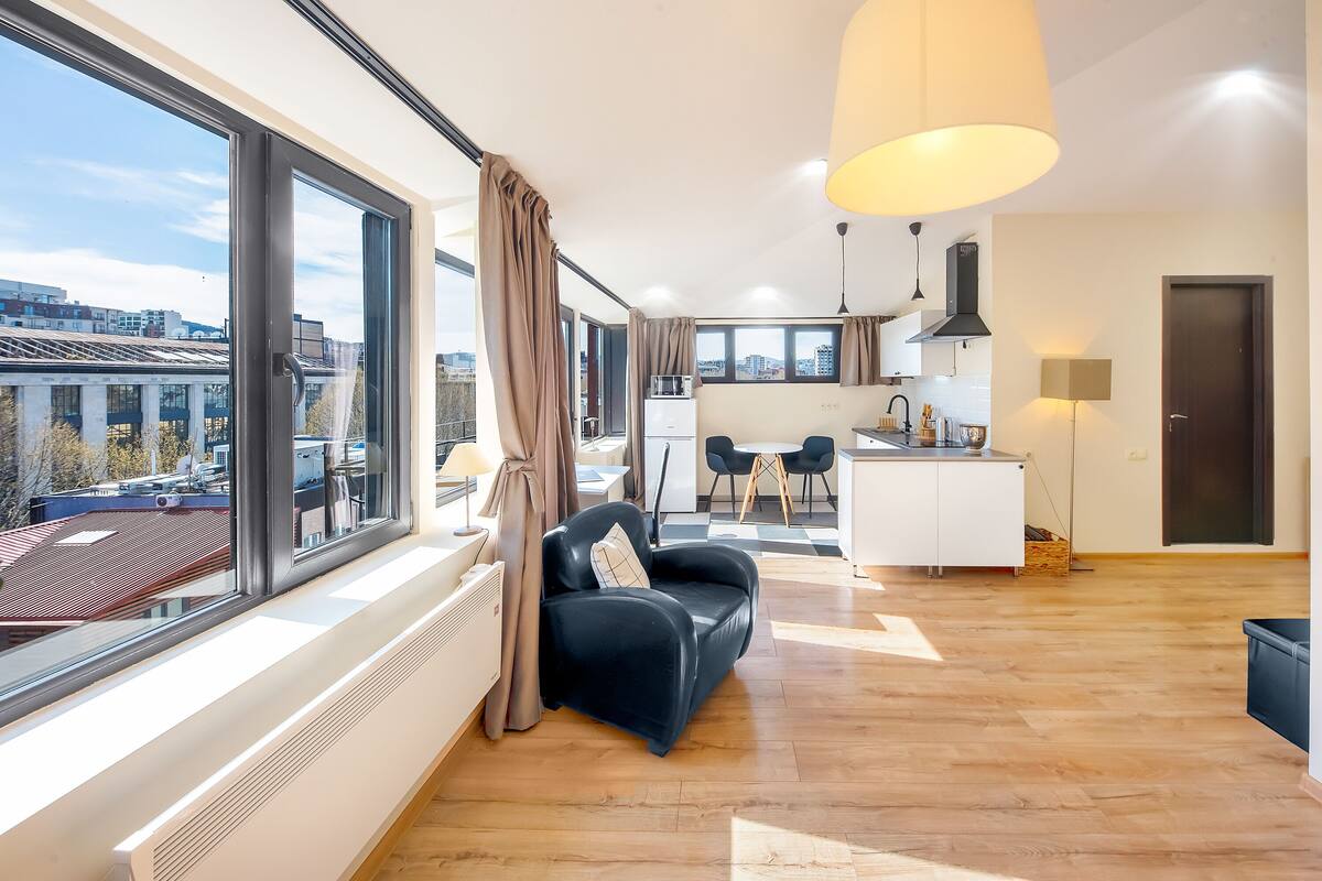 Eine moderne Wohnung mit großem Fenster, einem schwarzen Ledersofa, einer Küchenzeile und einem Esstisch.