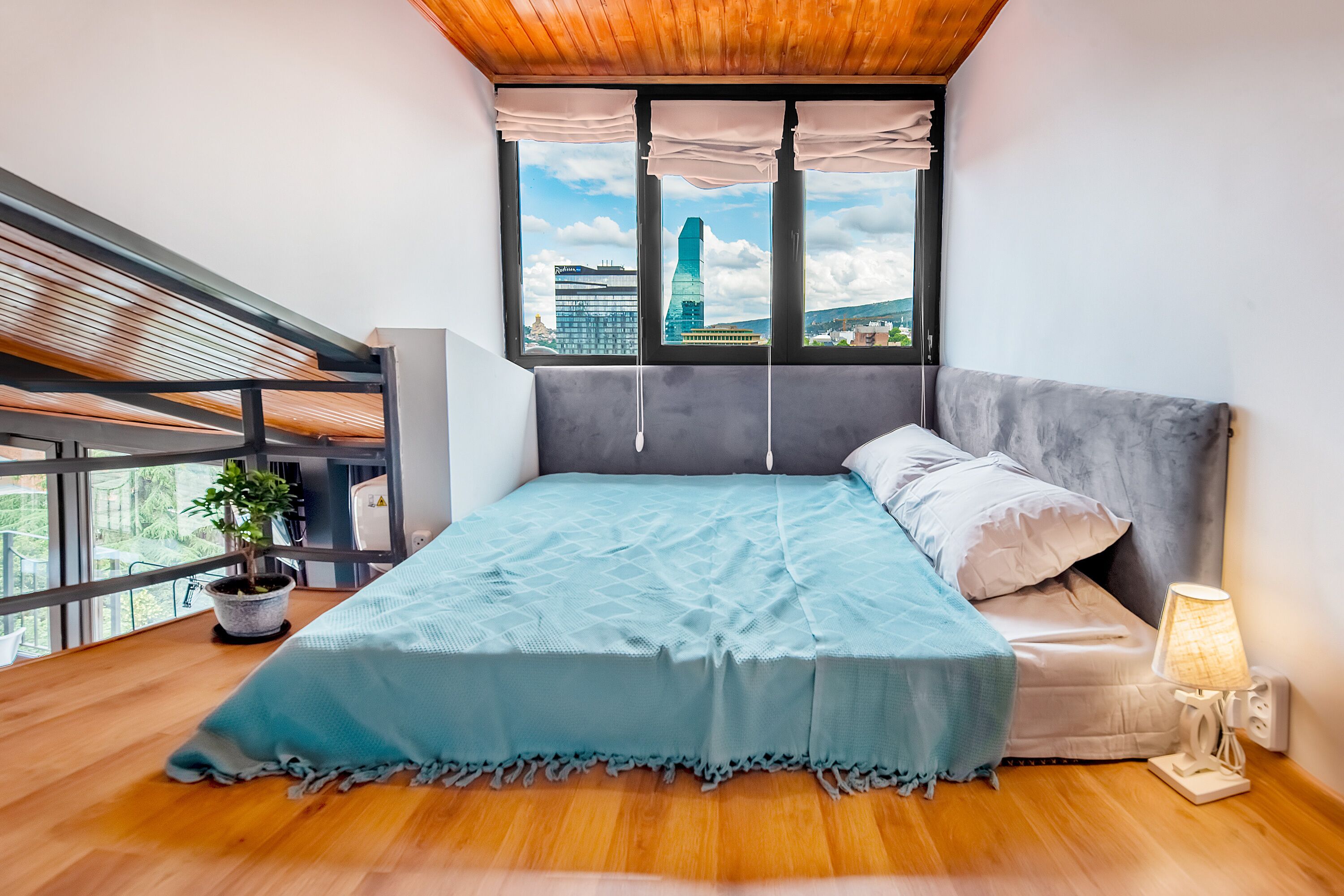 A bedroom with a bed, a potted plant, a lamp, and a view of a cityscape through a window.