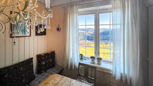 A bedroom with a large window, a wooden bed, a bedside table with a lamp, and a decorative chandelier. - 4 bedroom cozy home in Svorkmo (Svorkmo)