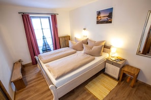 A bedroom with a bed, bedside lamps, a nightstand, a wooden bench, a window with curtains, and a framed picture on the wall.