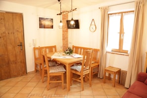A dining area with a wooden table and chairs, a small wooden side table, a clock on the wall, and a window with curtains. - Whg 8a Neubau, 60qm, 2 Schlafzimmer, Balkon (Eggstätt)