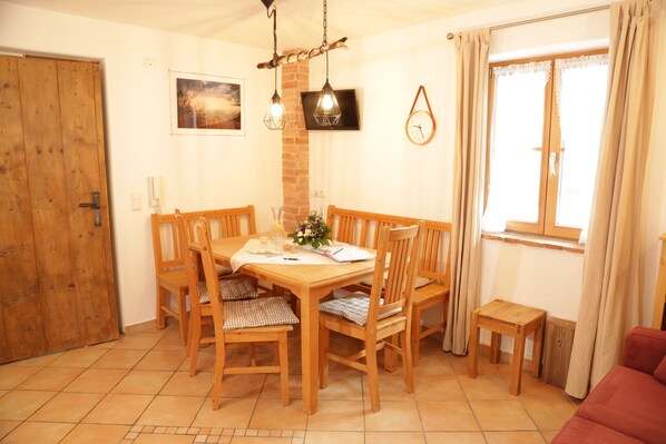 A dining area with a wooden table and chairs, a small wooden side table, a clock on the wall, and a window with curtains.