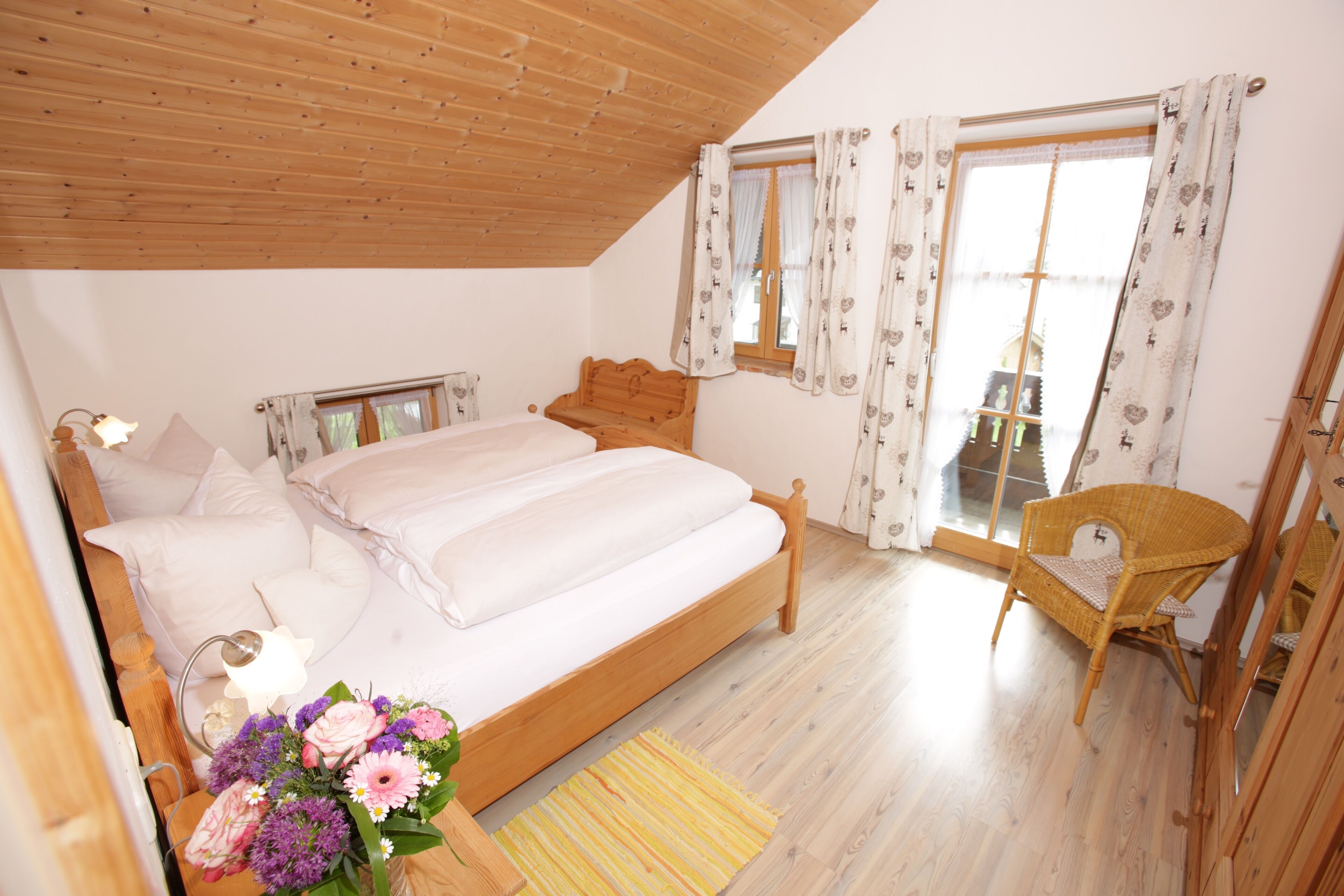 A bedroom with a wooden bed, a bedside table with a lamp, a chair, and a window with curtains.