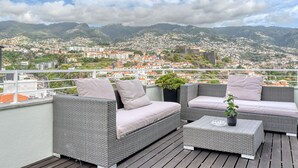 A balcony with wicker furniture and a potted plant, overlooking a town with mountains in the background.