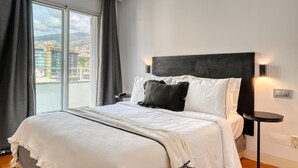 A hotel room with a large bed, a view of the city through the window, and a small side table.
