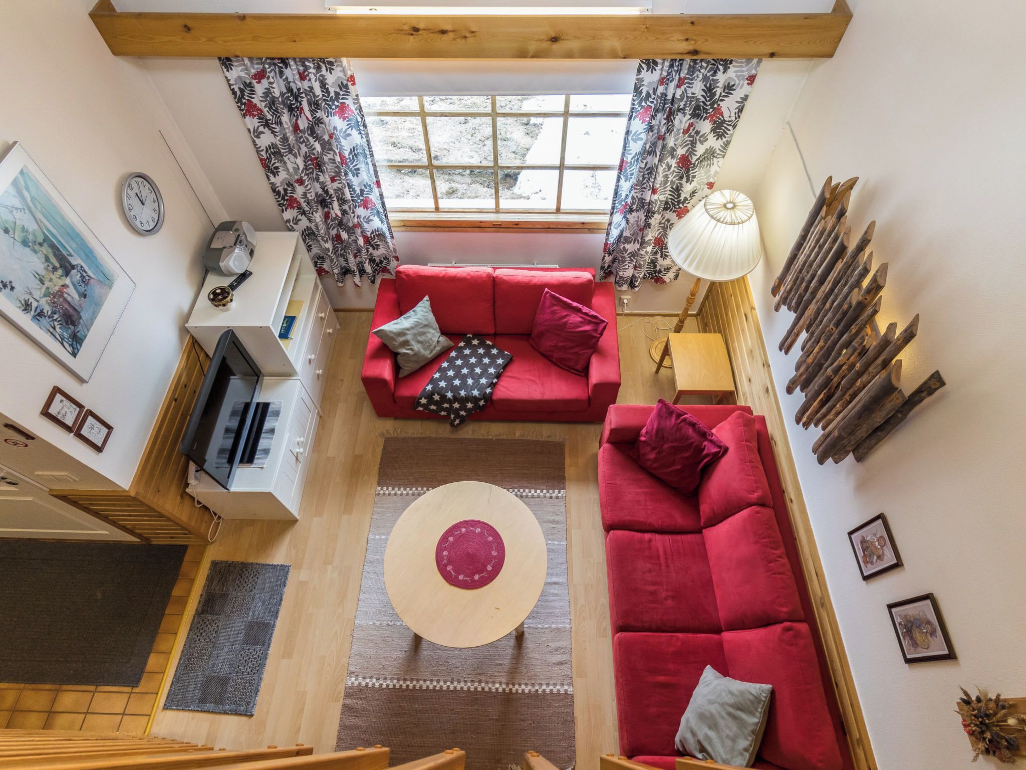 Un salon aux hauts plafonds, avec un canapé rouge, une table basse ronde et une vue sur l’extérieur à travers une fenêtre ornée de rideaux à motifs floraux.
