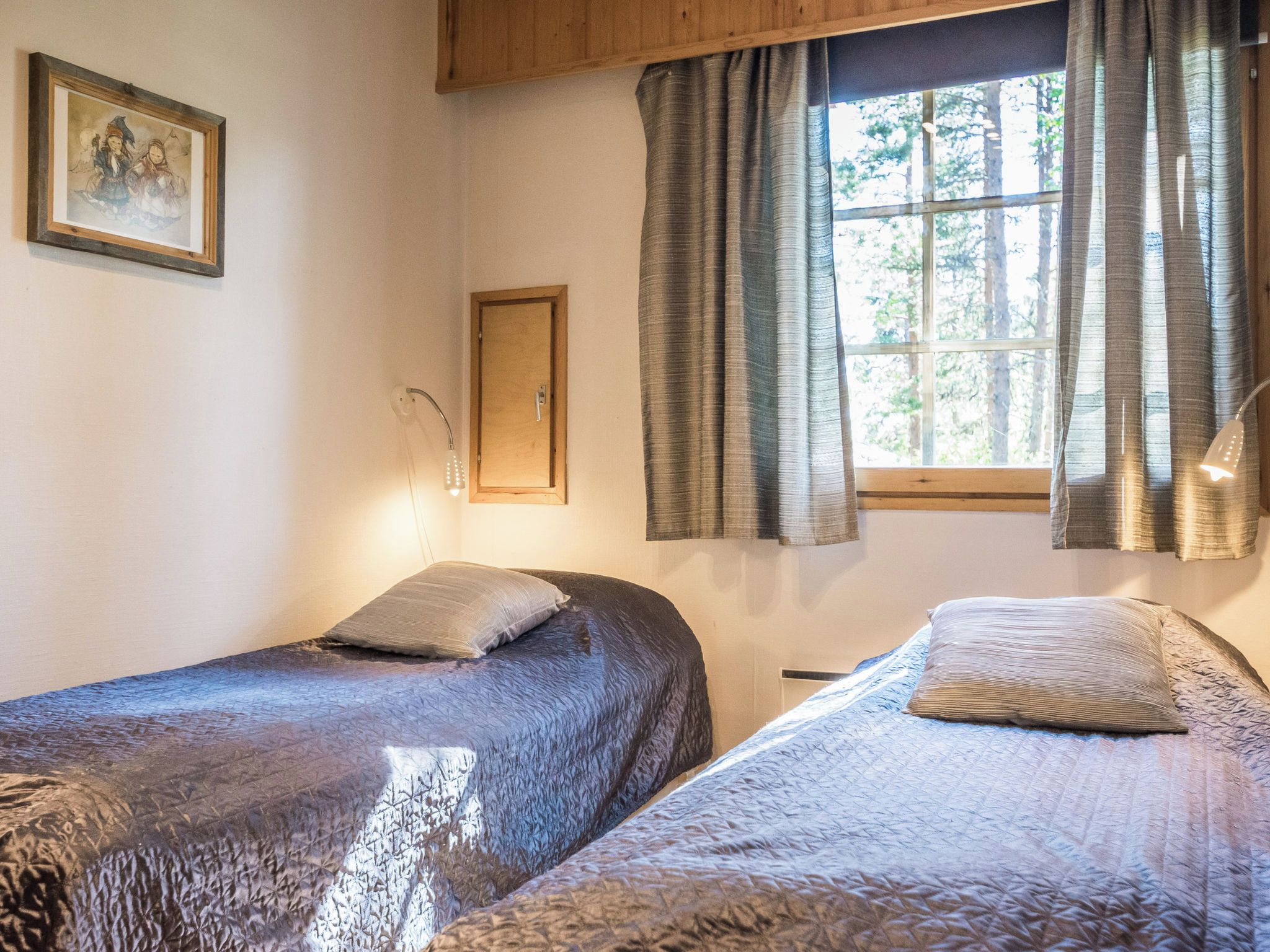 A room with two beds, a wooden ceiling, a window with curtains, and a wall-mounted picture.