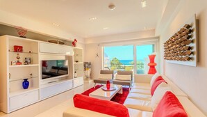 Una sala de estar moderna con un televisor de pantalla plana grande, sofás blancos, un sillón rojo y vistas al mar a través de un ventanal amplio.