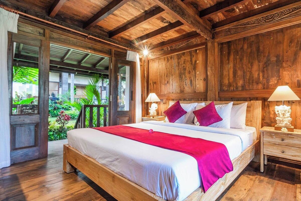 A wooden bedroom with a large bed, two bedside tables, and a view of greenery outside.