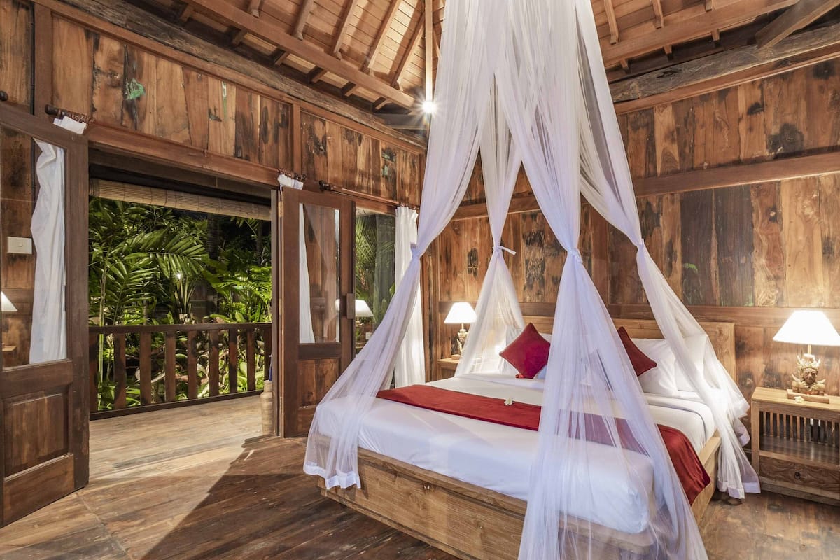 A wooden four-poster bed with white bedding and red accents, a nightstand with a lamp, and a view of lush greenery through an open door.