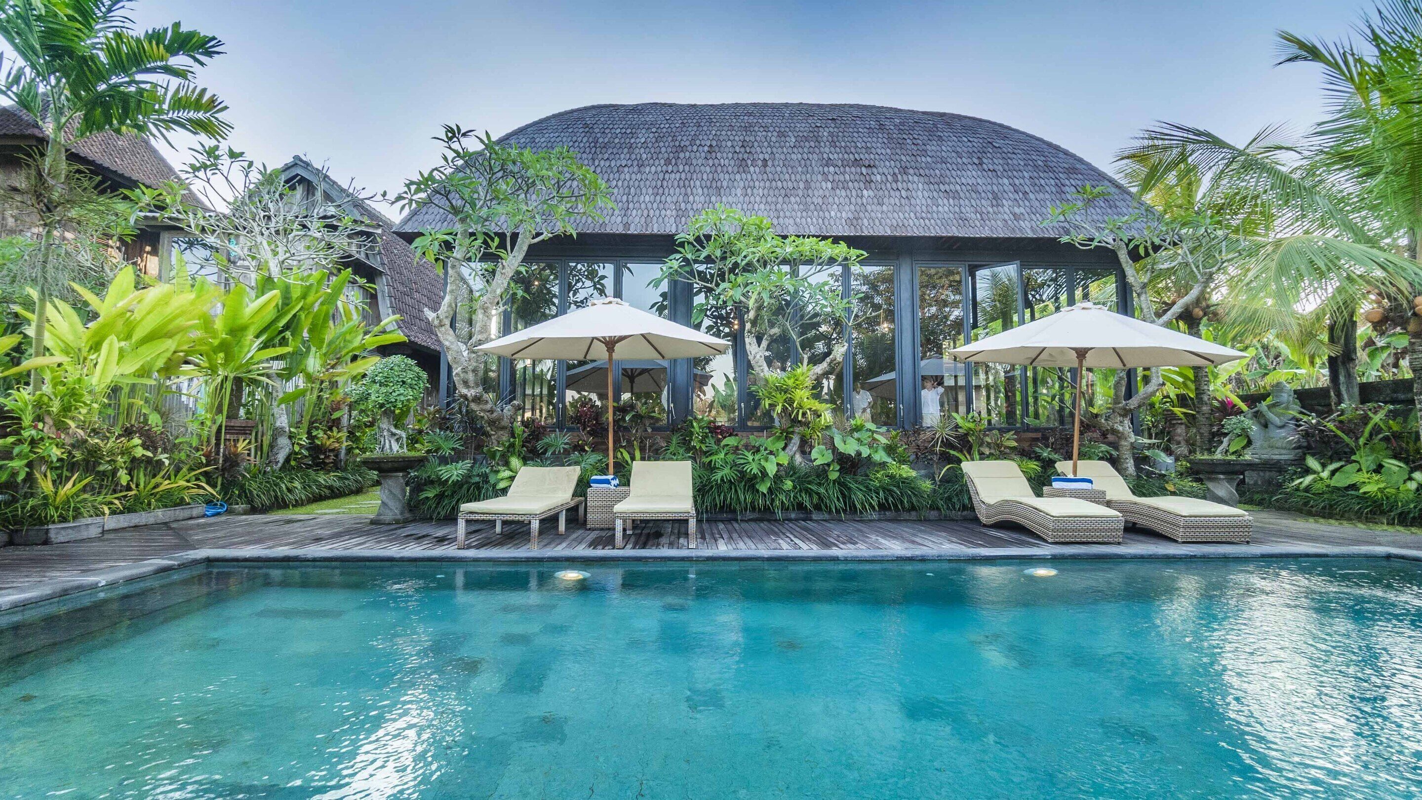 A pool area with lounge chairs and umbrellas, surrounded by lush greenery and a traditional building with a curved roof.