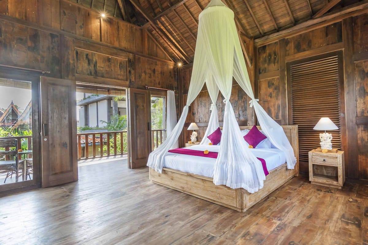 A wooden four-poster bed with a canopy, a nightstand with a lamp, and a view of the outdoor balcony.