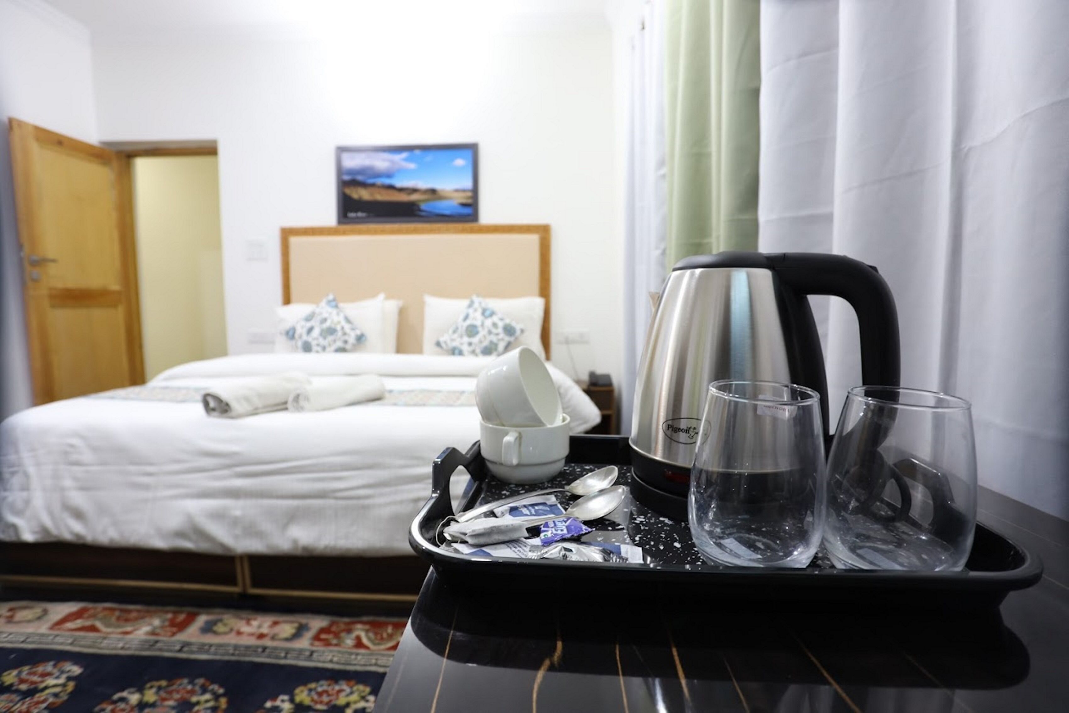 Quarto de hotel com cama, bandeja com copos e chaleira, e um quadro na parede.
