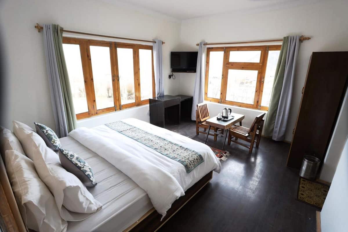 A bedroom with a large bed, a desk, and a chair. There are two windows with curtains, a TV mounted on the wall, and a wooden door.
