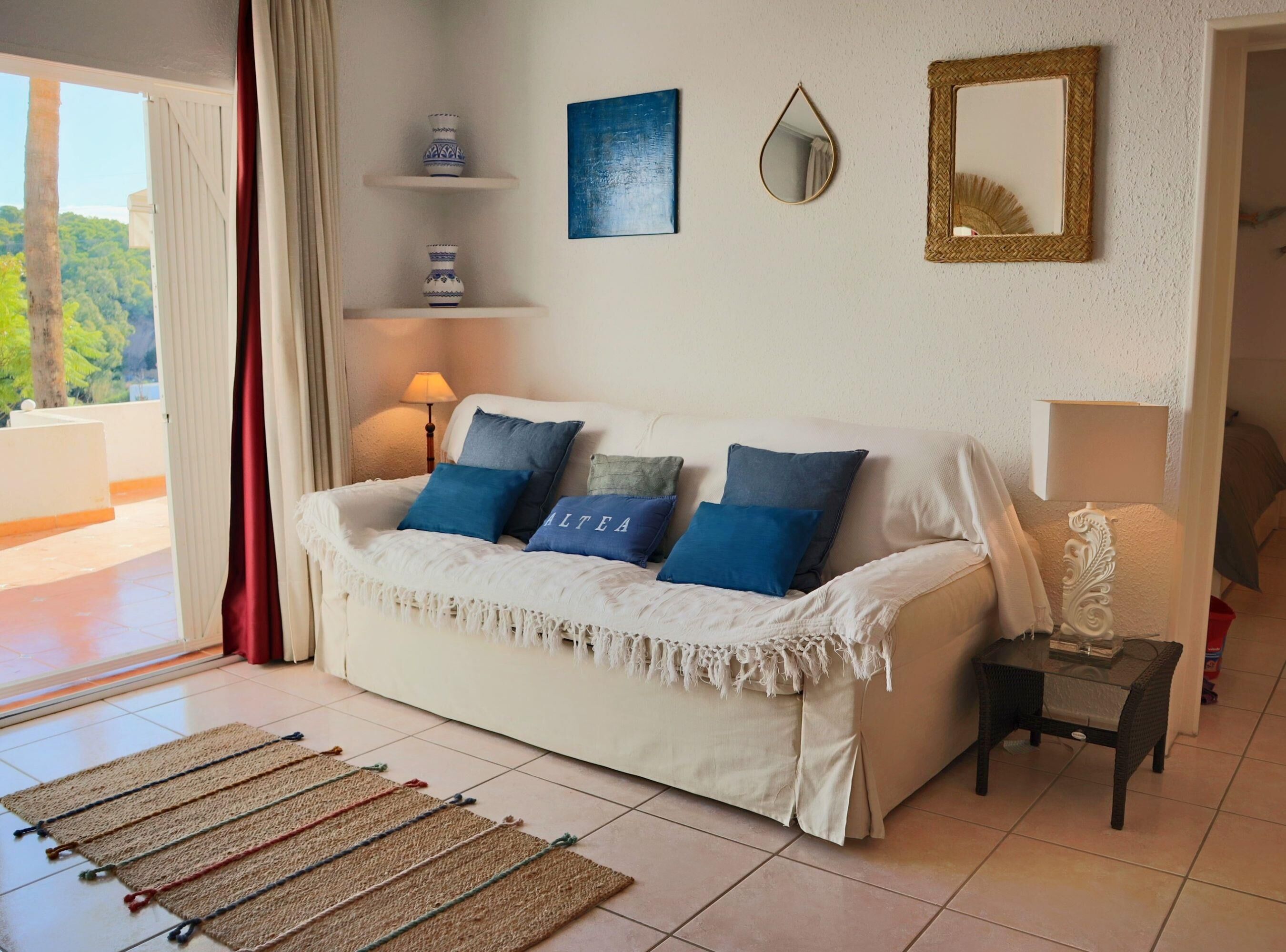 Een woonkamer met een witte bank, blauwe kussens, een bijzettafeltje met een lamp, en uitzicht op groen.