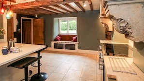 A kitchen with exposed wooden beams, a window with a view of greenery, a bench, and a stove. - Pentre Hall (Oswestry)
