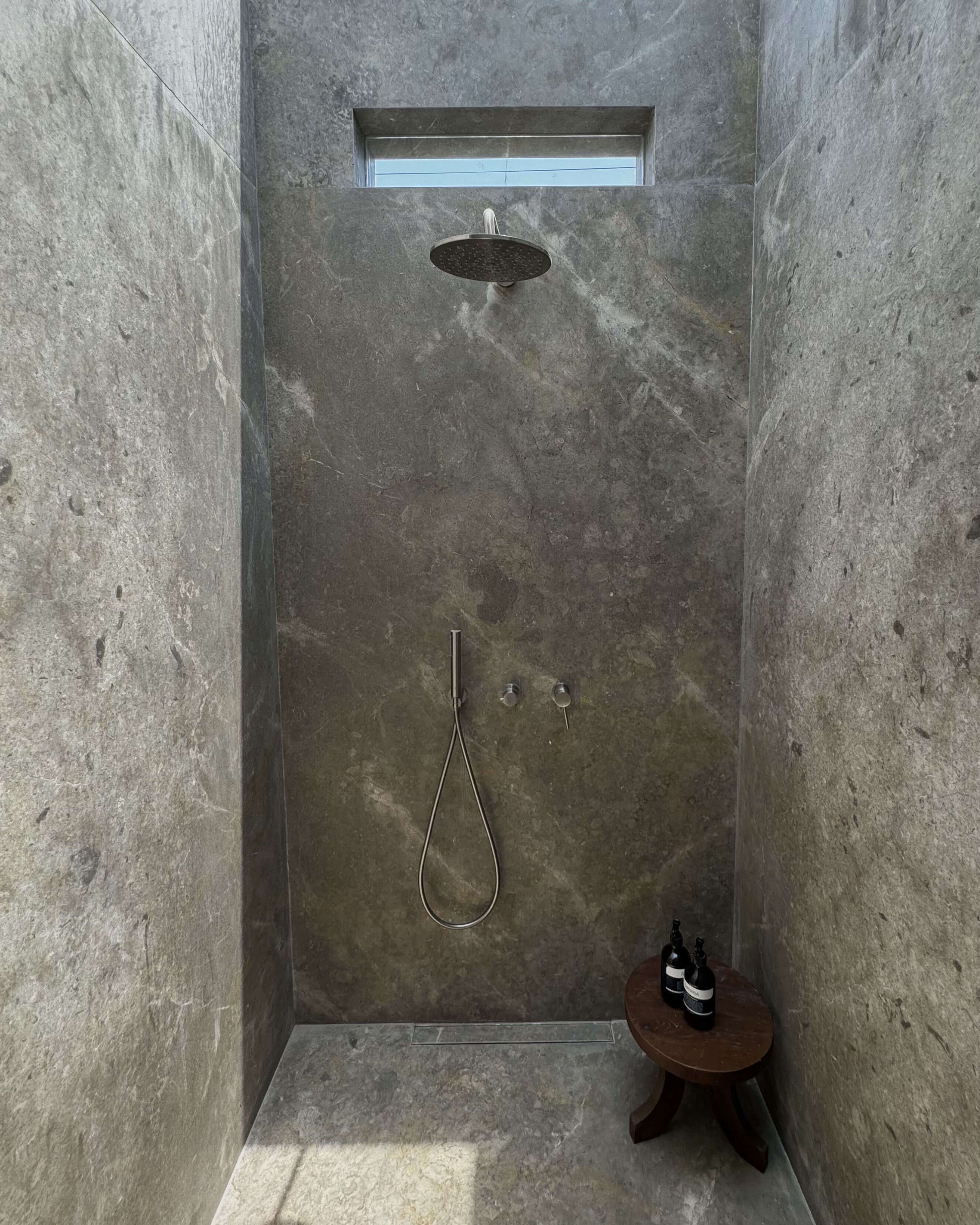 A modern shower with a rain showerhead and handheld showerhead, a small wooden stool, and a window above the shower.