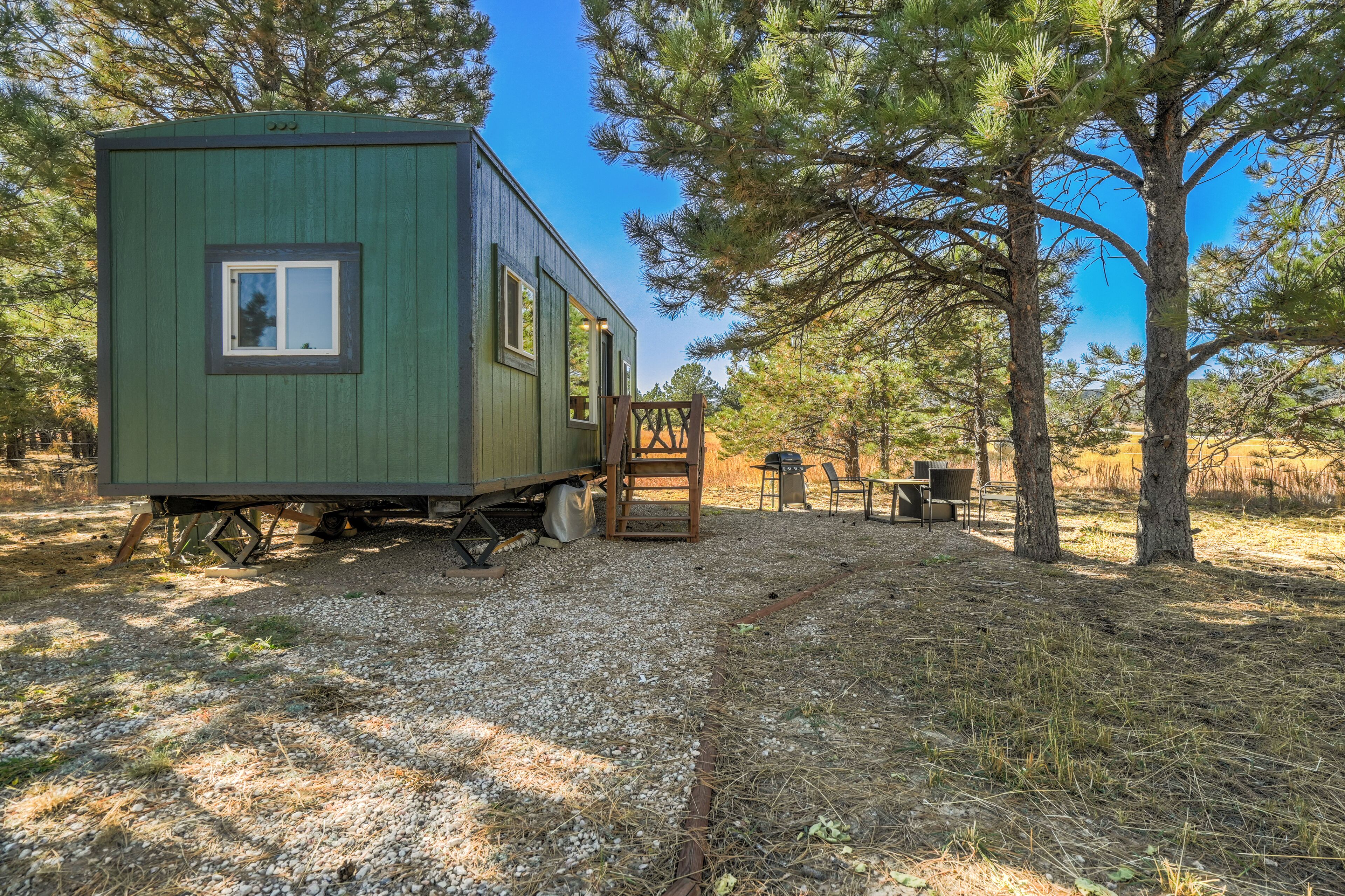 Une petite maison mobile verte, dotée d’une fenêtre et d’une porte, située dans un espace boisé, avec des arbres et un chemin de gravier.