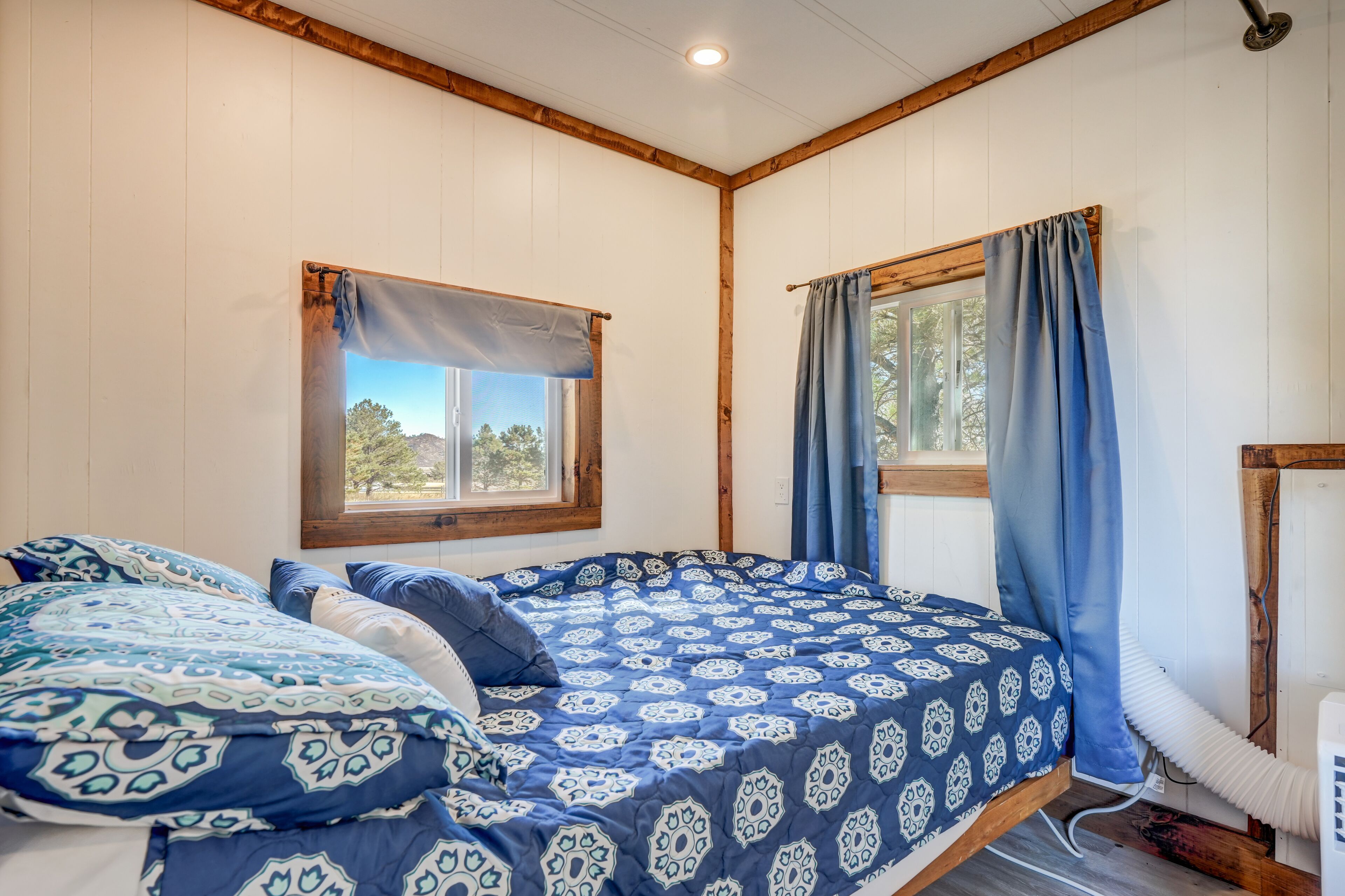 A bedroom with a bed, two windows with curtains, wooden beams on the ceiling, and a heater on the floor.
