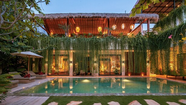 Un área junto a la piscina con una estructura de techo de paja, asientos interiores y exuberante vegetación.