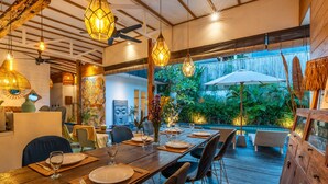 A wooden dining table set with plates, glasses, and cutlery, surrounded by chairs, under hanging lights, with a view of greenery and a white umbrella. - Private Pool • Hot Tub • Near Batu Bolong Beach (Canggu)