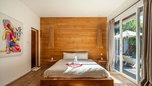 A modern bedroom with a wooden headboard, a large bed with a heart-shaped decoration, a painting on the wall, and a view of the outdoors through a sliding glass door. - Private Pool • Hot Tub • Near Batu Bolong Beach (Canggu)