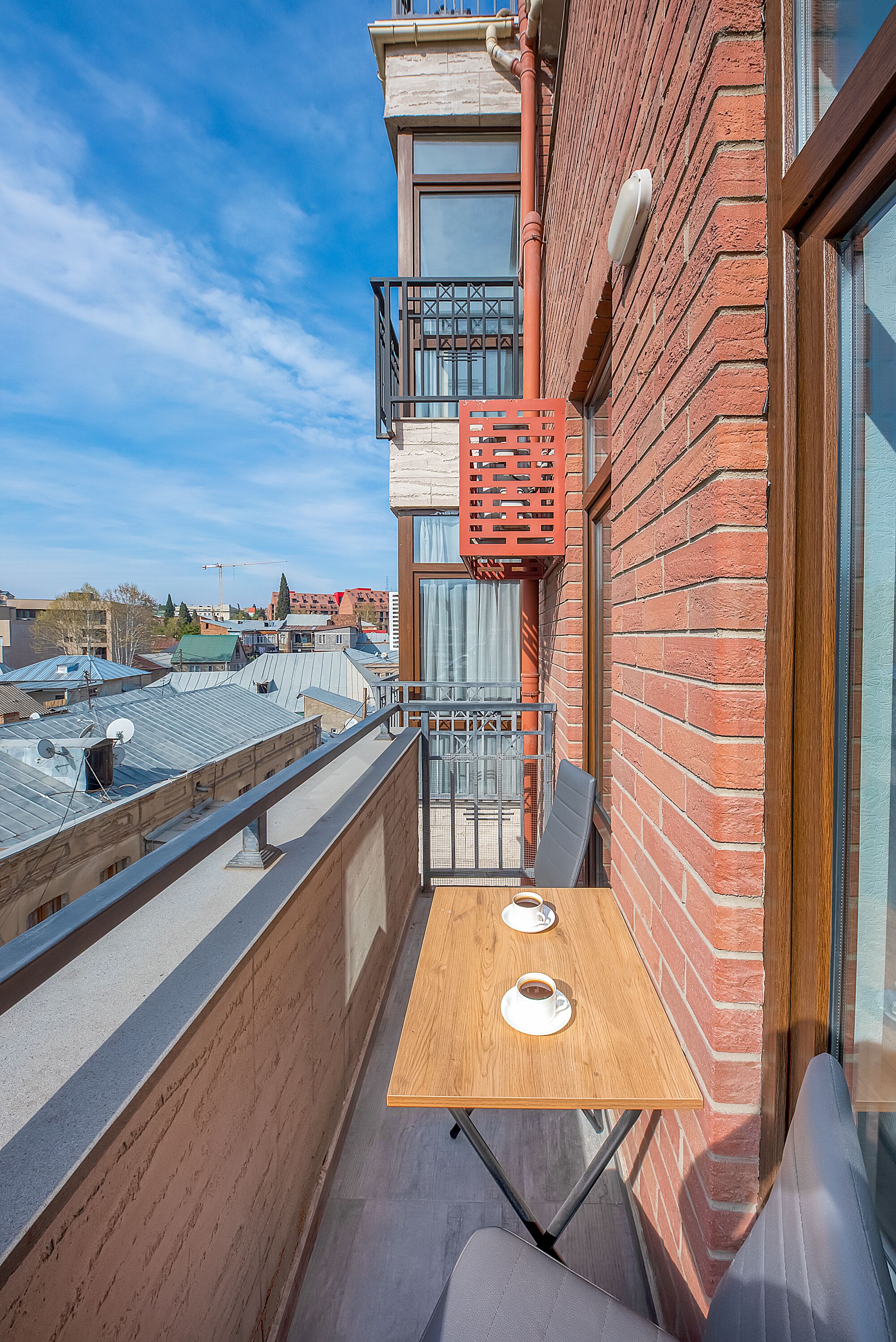 A balcony with a wooden table and two cups, a red brick wall, and a view of the cityscape.