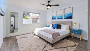 A bedroom with a bed, a ceiling fan, a window, and a nightstand with a lamp. - SoCal Private Pool Bungalow |4BR (Indio)