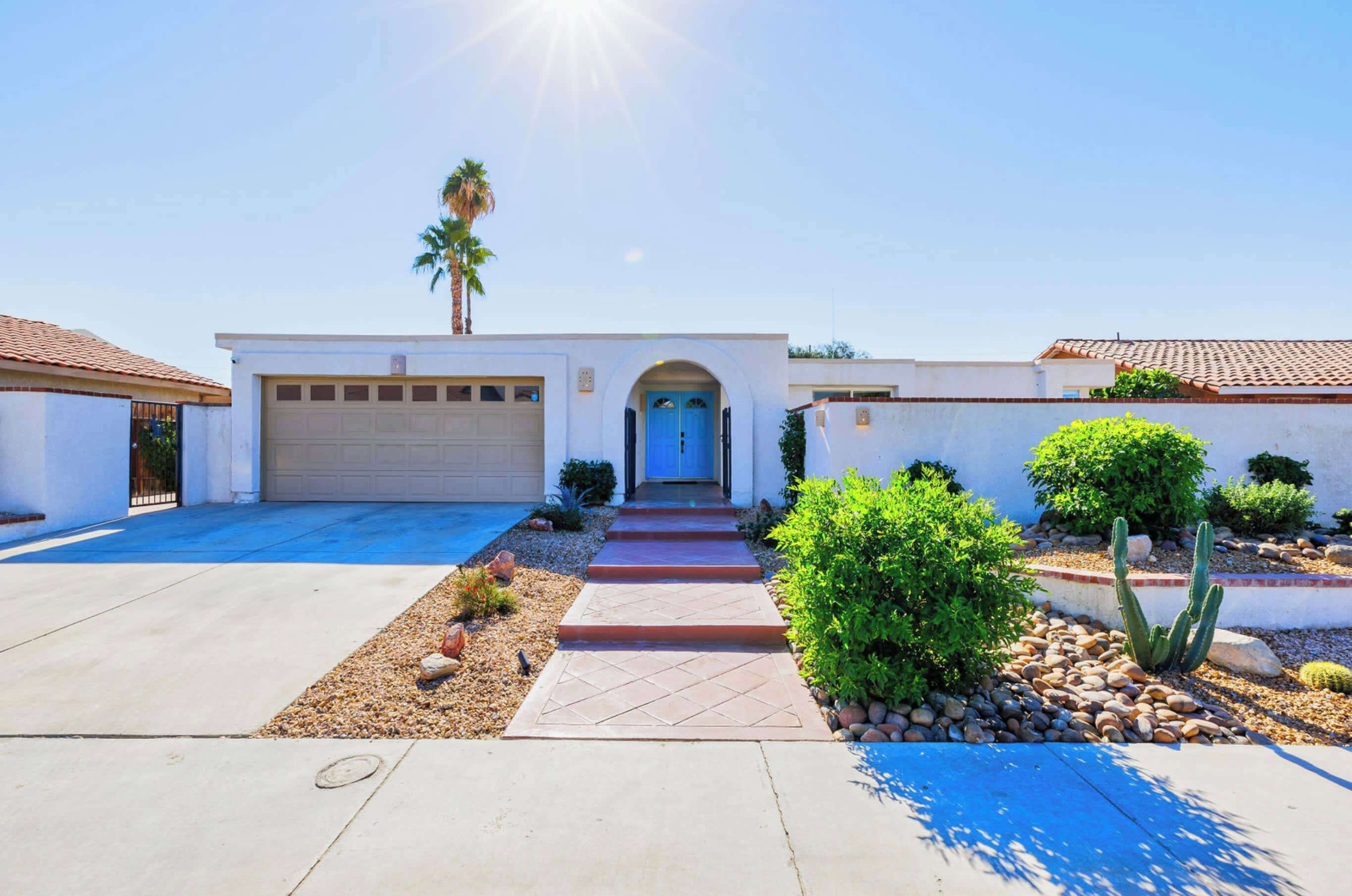 A residential house with a garage, a blue door, a pathway leading to the entrance, and a small garden with cacti and shrubs.
