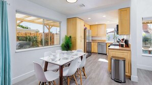 A modern kitchen with wooden cabinets, a stainless steel refrigerator, and a dining area with white chairs and a table. - SoCal Private Pool Bungalow |4BR (Indio)