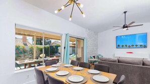 A dining area with a table set for six, a grey sofa, a ceiling fan, and a TV mounted on the wall. - SoCal Private Pool Bungalow |4BR (Indio)