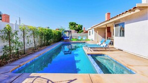 A swimming pool with lounge chairs, a small structure, and a white building with a red roof. - SoCal Private Pool Bungalow |4BR (Indio)