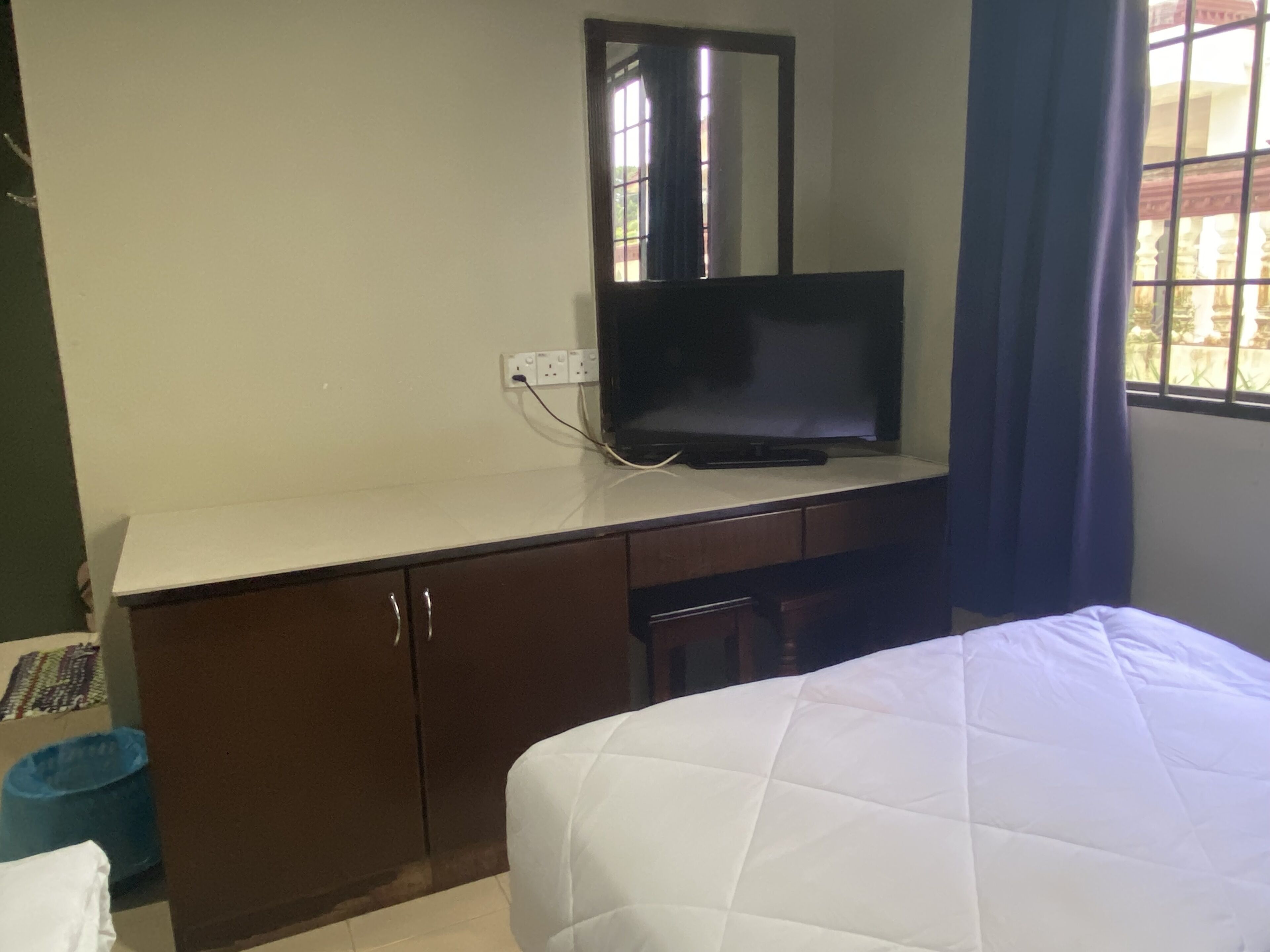 Quarto de hotel com cama, televisão sobre uma cômoda, janela com cortinas e lixeira.
