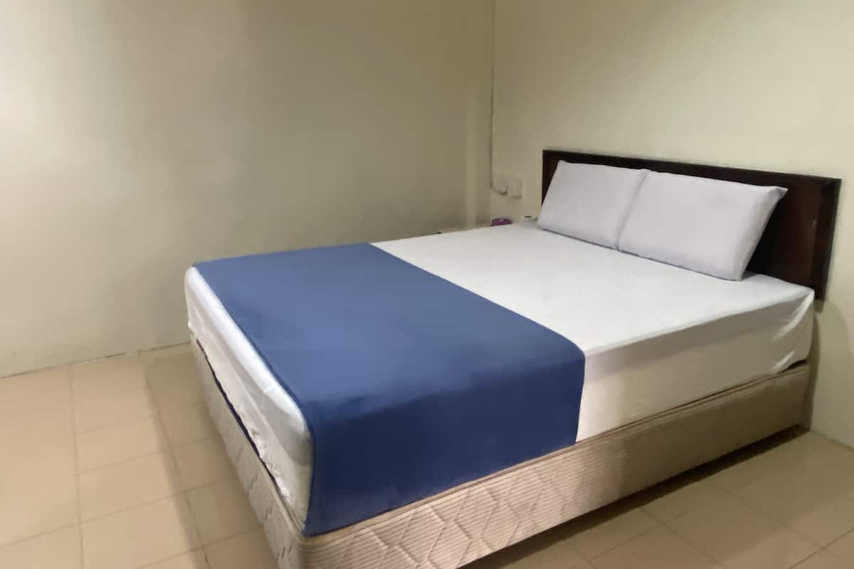 Un lit simple avec une couvre-lit bleue et des oreillers blancs, une tête de lit en bois et un mur uni avec un interrupteur.
