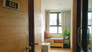 A hallway with a wooden door labeled '501', a bench with a cushion, a small table with a plant, and a window with curtains. - BM Loft On Nut & Massage (Bangkok)