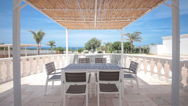 An outdoor patio with a wooden pergola, white chairs, and a table.