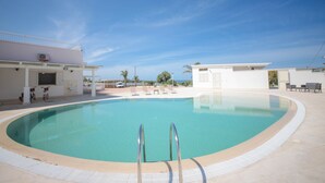 A clear blue swimming pool with a metal ladder, surrounded by lounge chairs and a white building with a balcony.