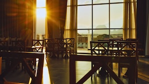 A room with wooden chairs arranged in rows, large windows with curtains, and a view of mountains. - Oudomsin VangVieng Hotel (Vang Vieng)