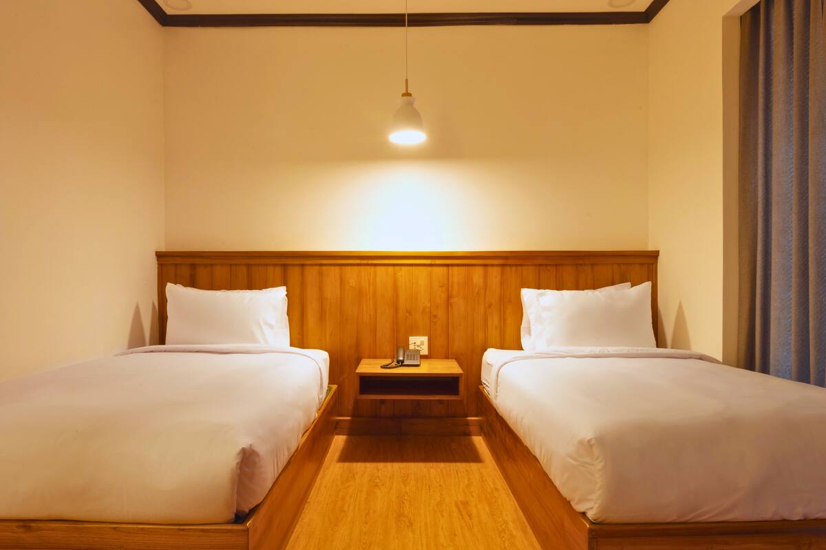 Een hotelkamer met twee eenpersoonsbedden, een houten hoofdbord, een nachtkastje met lamp en een raam met gordijnen.