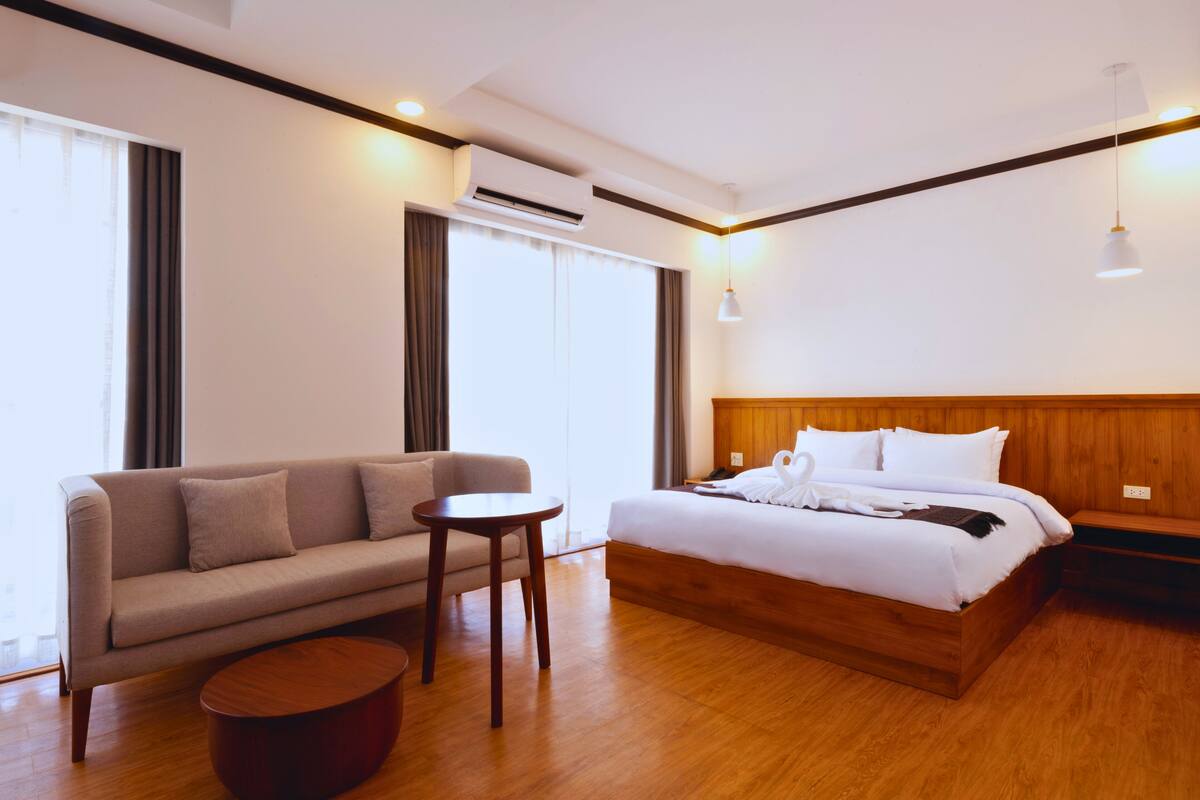 Een hotelkamer met een houten bed, een bank, een klein rond tafeltje en een groot raam met gordijnen.