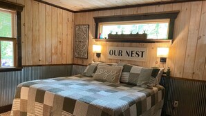 Une chambre confortable à l’ambiance chaleureuse, avec un décor en bois, comprenant un lit recouvert d’une courtepointe, une inscription « NOTRE NID » au-dessus, et une fenêtre ornée de plantes.