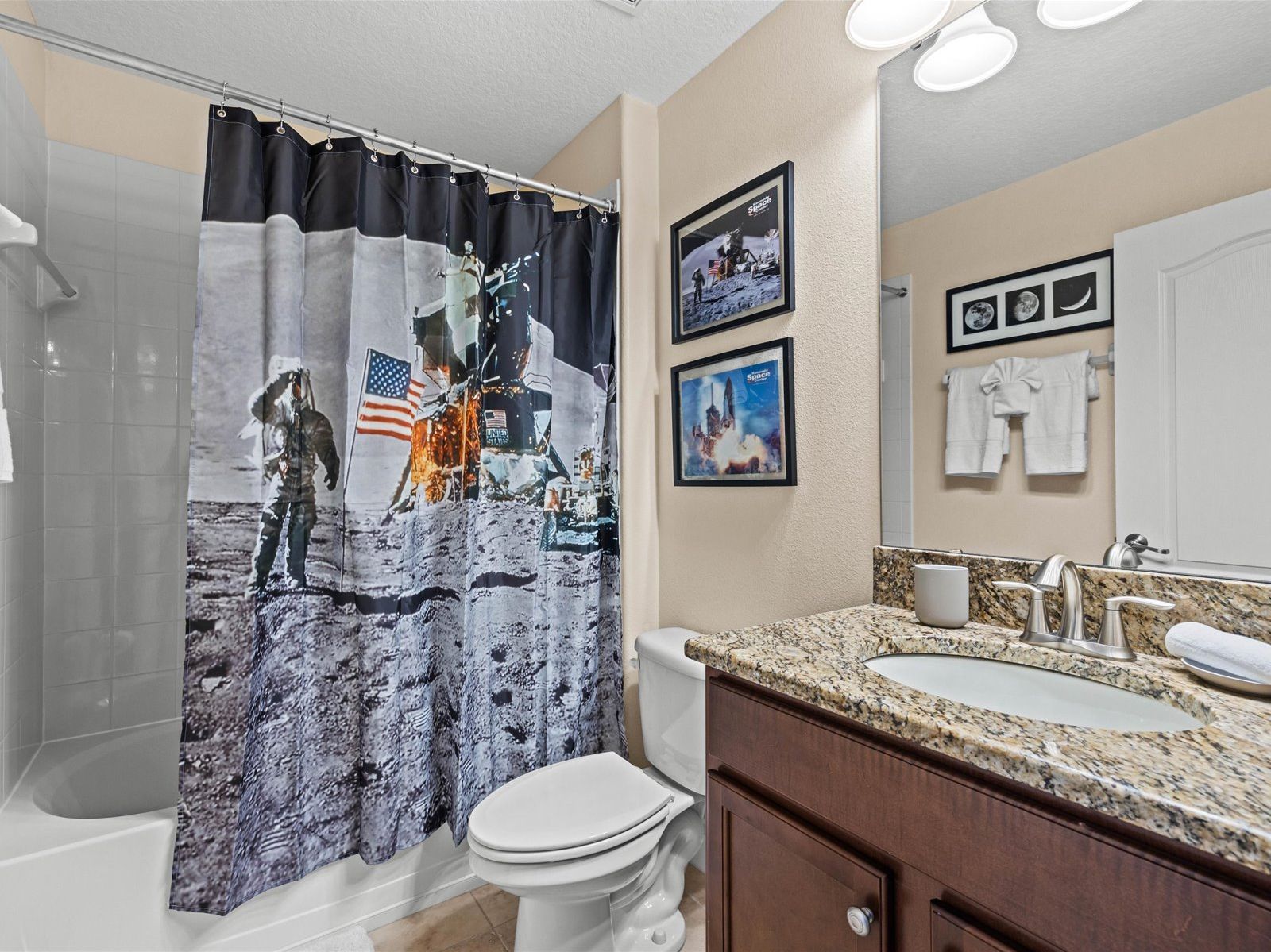 A bathroom with a shower curtain depicting a space shuttle launch, a white toilet, a granite countertop sink, and a mirror with framed pictures of the moon and planets.