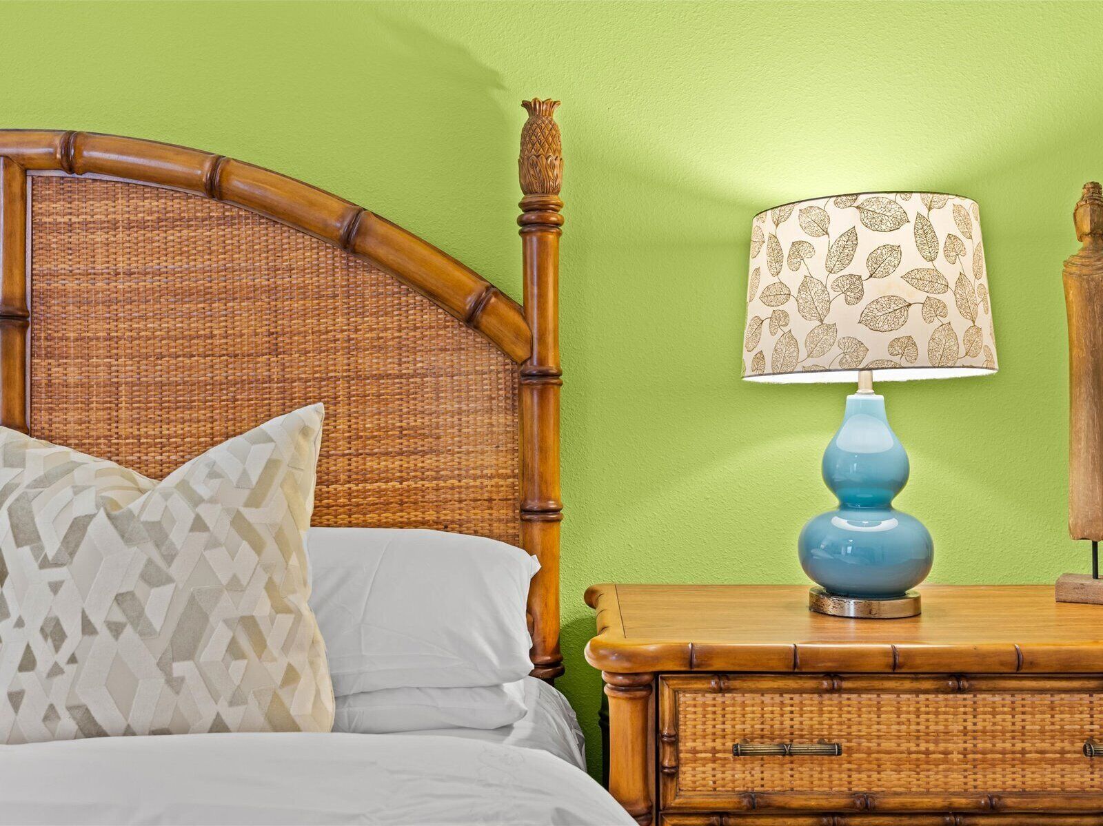 Een bed met een geweven hoofdbord, een kussen met geometrisch patroon en een blauwe lamp met een bladmotief aan de zijkant op de muur.