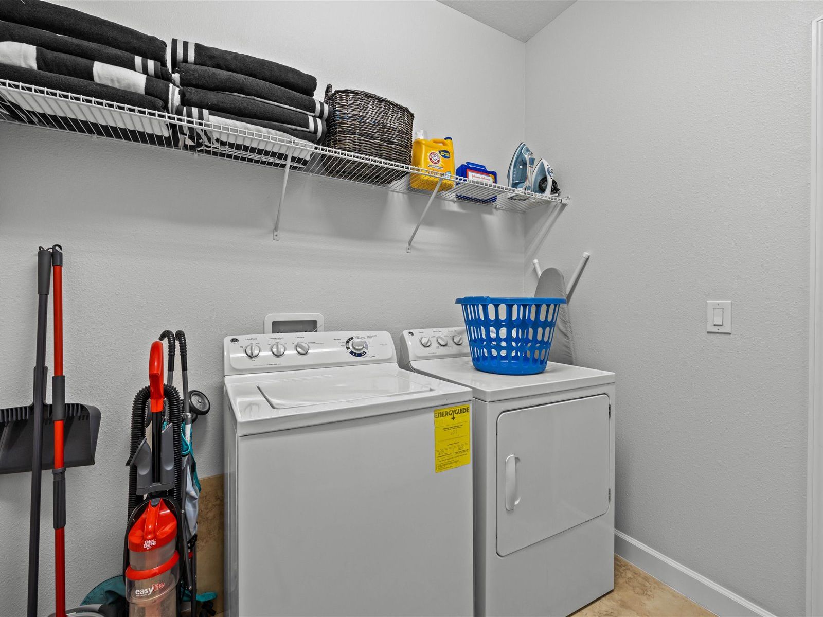 A laundry room with a front-loading washer and dryer, a mop, a broom, and a laundry basket on the dryer.