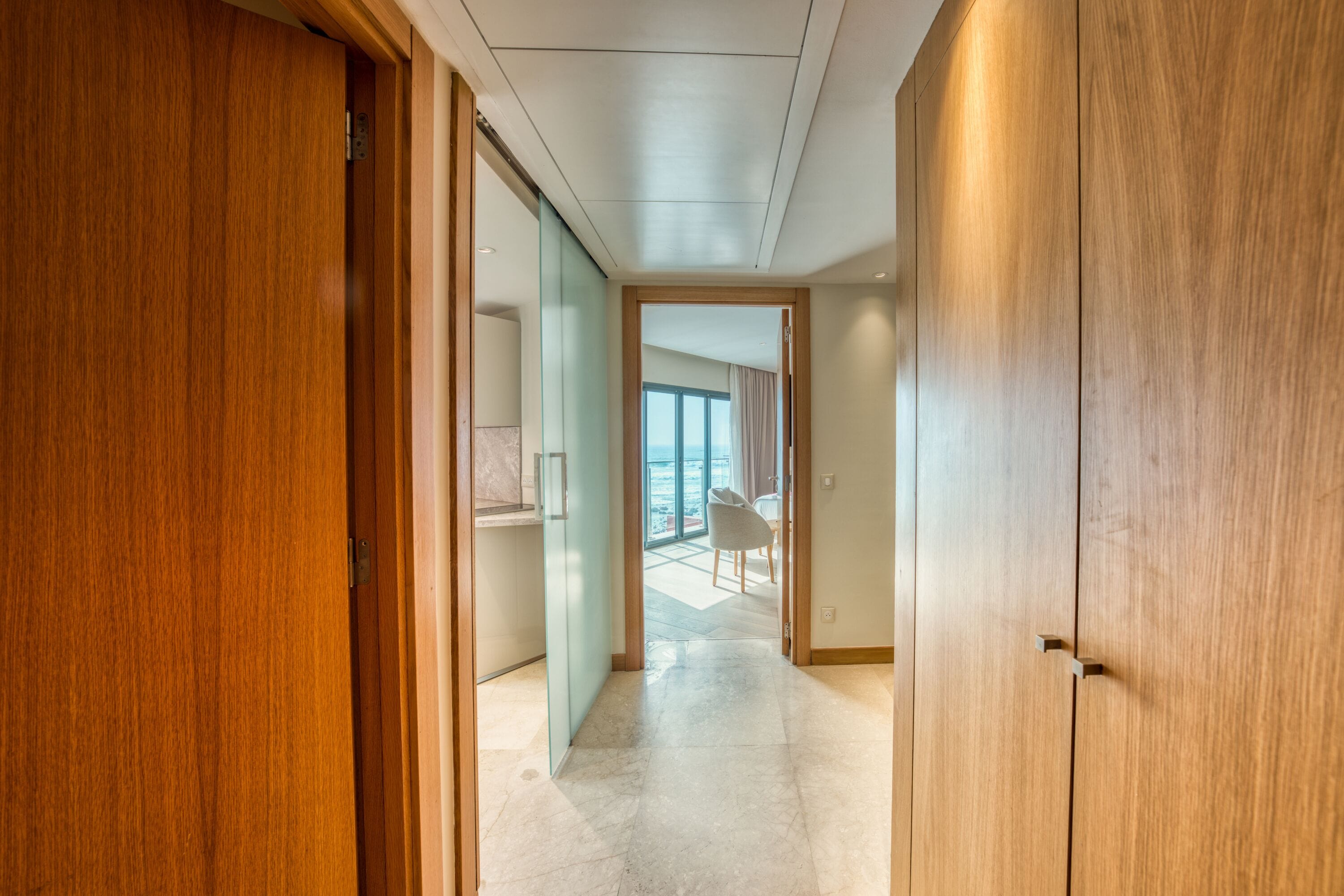 Passadís amb portes de fusta i vista a una habitació amb una finestra gran i una cadira.